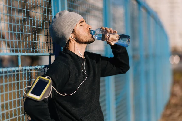 Young male runner drinking water from a bottle while leaning on a fence outdoors.