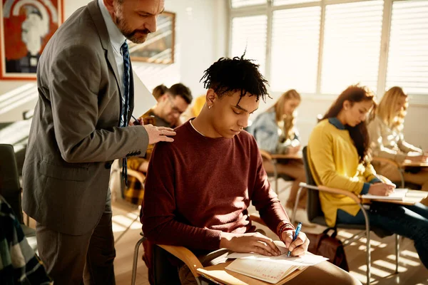 College teacher monitoring black male student who is writing exam in the classroom.