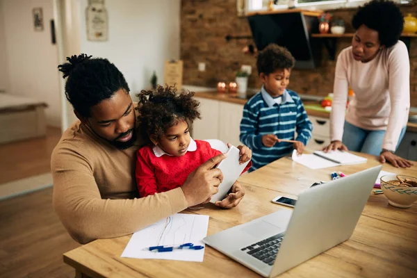 African American father working on touchpad and laptop while daughter is sitting on his lap at home. 