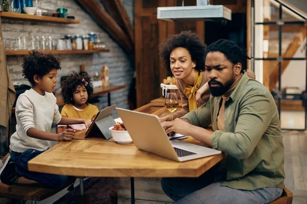 Black parents working on laptop while their kids are surfing the net on touchpad at home.