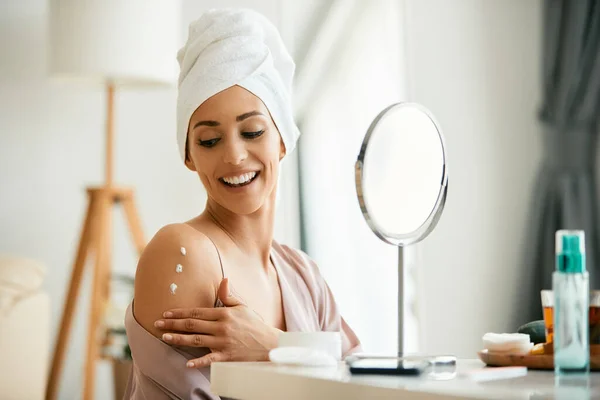 Young happy woman applying body cream on her arms and shoulder after taking a bath at home.
