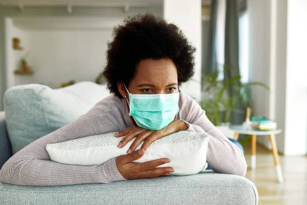Black woman with face mask feeling bored and thinking of something while lying down on sofa at home during virus epidemic.