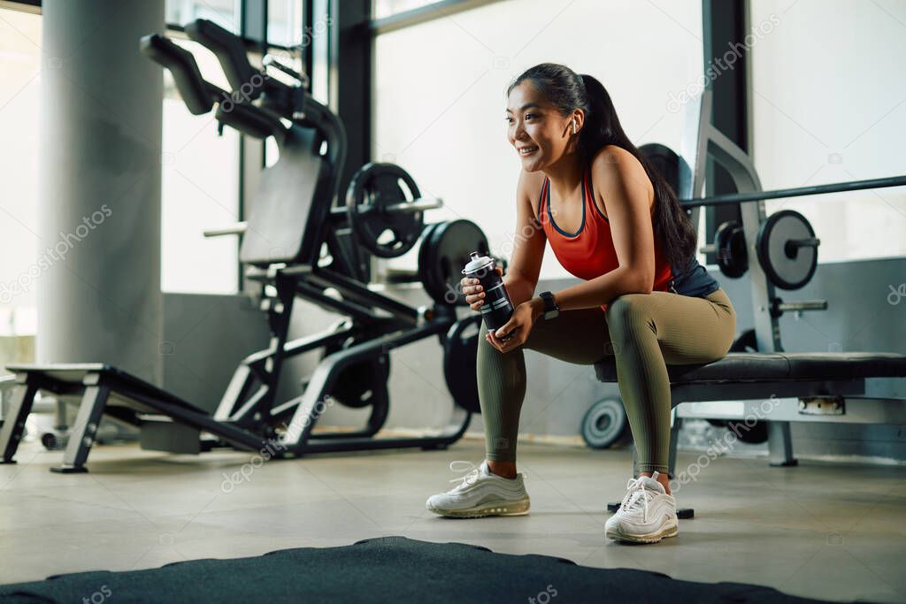 Happy Asian sportswoman having a water break while working out in a gym. 