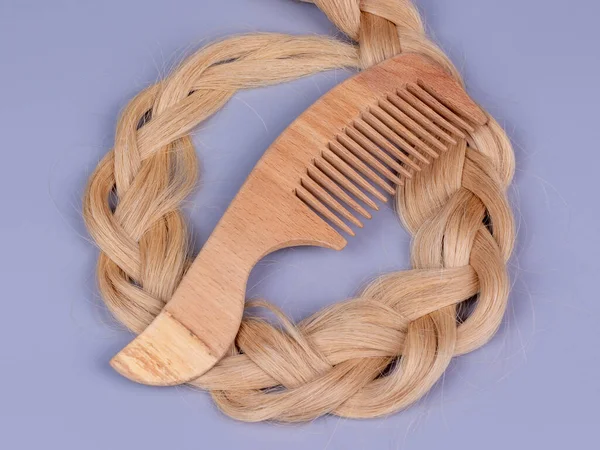 A wooden comb lies on the hair in the form of a light-colored braid, on a gray background. The concept of preserving naturalness, hair care, donation. Long natural hair. Tools from ecological material