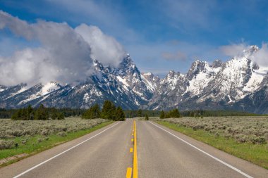 Highway pointed toward the high, snow-capped peaks of the Teton Range under blue sky with dramatic clouds in Grand Teton National Park, Wyoming