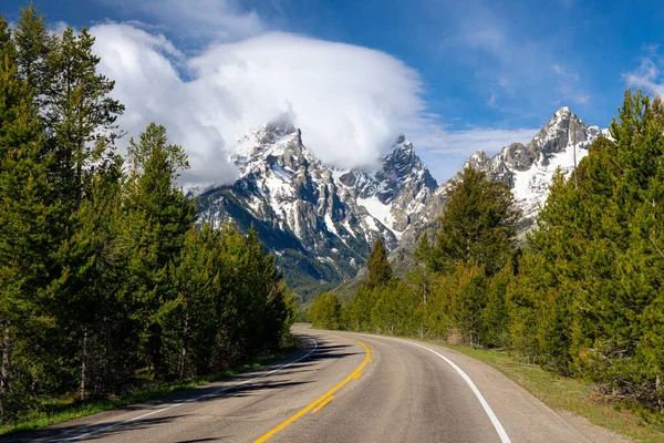 Highway curves towards snow-capped mountain peaks of the Teton Range under dramatic clouds and blue sky in Grand Teton National Park, Wyoming