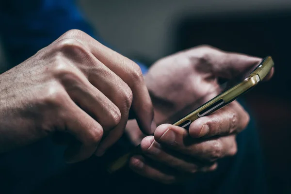 İki eliyle akıllı telefonu tutan ve kullanan bir adam. Yakın plan, koyu ton, bulanık arkaplan.