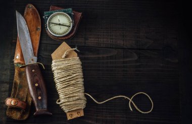 Old rusty hunting bushcraft knife, military compass and a linen rope on the wooden table. Leather cases, copy space, top view, survival concept.