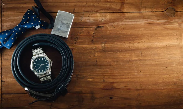 Bowtie, silver wristwatch, black leather belt and old chrome lighter on the wooden table. Top view, male accessories, elegance concept, copy space.