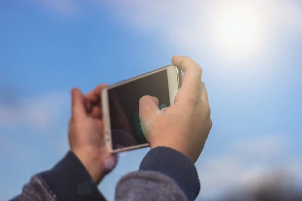 Little boy or a girl holding a smartphone using both hands and taking a photo of the blue sky. Sunny outdoors background, travel concept, close up.