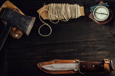 Old rusty hunting bushcraft knife, military compass, small dusty axe and a linen rope on the dark wooden table. Leather cases, copy space, top view, survival concept.