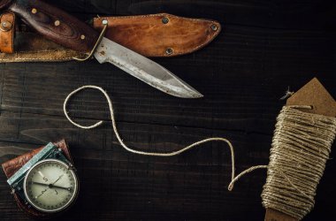 Old rusty hunting bushcraft knife, military compass and a linen rope on the wooden table. Leather cases, top view, survival concept.