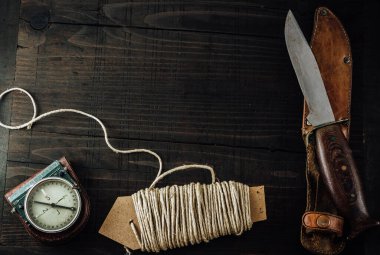 Old rusty hunting bushcraft knife, military compass and a linen rope on the wooden table. Leather cases, copy space, top view, survival concept.