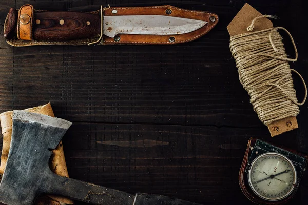 Old rusty hunting bushcraft knife, military compass, small dusty axe and a linen rope on the dark wooden table. Leather cases, copy space, top view, survival concept.