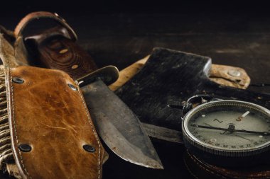 Old military compass, rusty hunting bushcraft knife, and a small axe on the dark wooden table. Leather cases, front view, survival hunting concept.