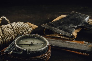 Old military compass, rusty hunting bushcraft knife, small axe and a linen rope on the dark wooden table. Leather cases, front view, survival hunting concept.