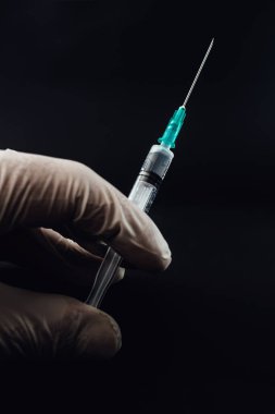 Close up of hand holding syringe. Doctor or nurse wearing rubber gloves and holding a syringe with attached needle. Covid-19 corona vaccine, medical healthcare and treatment concept, black background.