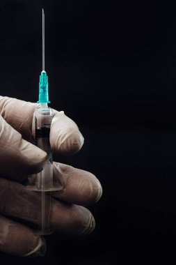Close up of hand holding syringe. Doctor or nurse wearing rubber gloves and holding a syringe with attached needle. Covid-19 corona vaccine, medical healthcare and treatment concept, black background.