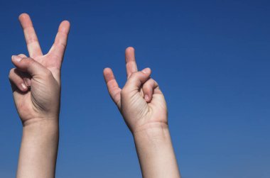Children's hands showing peace victory sign. Both hands raised on the clear blue sky. Gradient background, copy empty space for text, body parts concept.