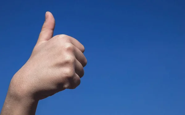 Children\'s hand showing thumbs up sign, a symbol of approval. Kid\'s hand raised against the clear blue sky. Close up, gradient background, copy empty space for text, body parts concept.