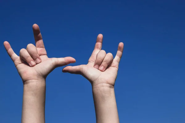 Children\'s hands showing devil horns sign, symbol of heavy metal and rock music. Both hands raised on the clear blue sky. Gradient background, copy empty space for text, body parts concept.