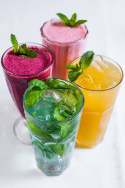 Çeşitli dört içki: ahududulu smoothie, çilekli smoothie, klasik limonata, portakallı limonata. İçkiler uzun şeffaf bardaklarda, üzerine nane ve fesleğen serpintileri. Gözlükler beyaz arka planda durur.