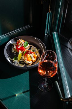Sandwiches with indal, lemon, mint, apricot, pickled pepper and white sauce in a black plate against the background of a dark green window with a glass of wine
