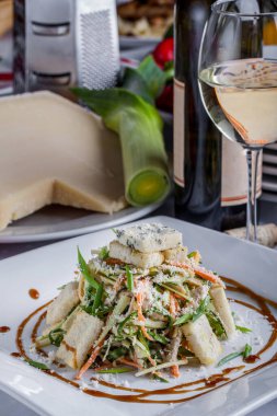 Tabakta Sezar salatası, bir bardak şarap, büyük bir parça peynir ve arka planda pırasa.