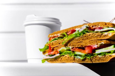 Sandwich in white paper with ham, lettuce, tomatoes, cucumbers and sauce with a paper cup of espresso with a lid standing on a white chair against the background of a white wooden wall
