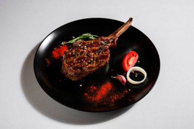 Fried pork steak on the bone with peppercorns, tomatoes, onions, paprika, salt, rosemary and garlic in a plate on a white background