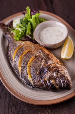 Grilled dorado stuffed with lemon slices with white sauce and arugula salad on an oval plate and a wooden table. Horizontal orientation 