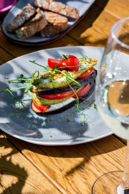 Eggplant, zucchini, tomatoes, pea sprouts, white sauce, pistachios in a plate on a wooden table with a glass of wine and bread in a plate