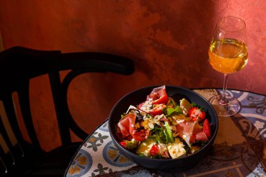 Salad with prosciutto, strawberries, mango, cheese, almond slices, arugula, salad and sauce in a plate on the table with a glass of wine against a brown wall