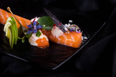 Dantel parçalı çiğ somon şeridi, havyar, çiçek yaprakları, siyah tabakta sos ve limon ve koyu bir arka plan. Yatay yönelim