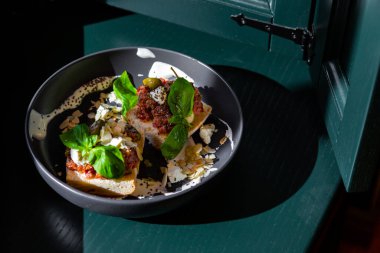 Sandwiches with cheese, meat, basil, almonds, kumquat, sauce and sesame seeds in a black plate against a dark green window background