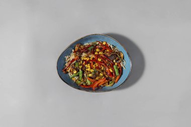 Tavada pişmiş sebzeli ev yapımı şehriye. Ev yapımı erişteler mavi bir seramik kasede. Mısır, kırmızı biber, yeşil fasulye, şampiyon mantarlar. Teriyaki soslu tüm sebzeler. Tabak gri bir arka planda duruyor..