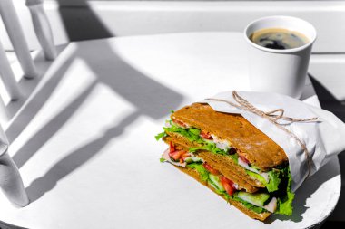 Sandwich in white paper with ham, lettuce, tomatoes, cucumbers and sauce with cappuccino with a paper cup standing on a white chair against the background of a white wooden wall