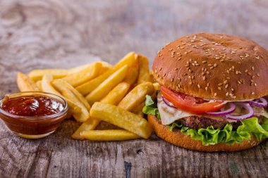 Burger with sesame bun, cutlet, tomatoes, onions, lettuce, cheese and mayonnaise with fries and sauce on a wooden table. Horizontal orientation