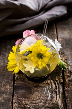 Cocktail with flowers and a straw in a glass which stands on a wooden table and a cloth and flowers on the background