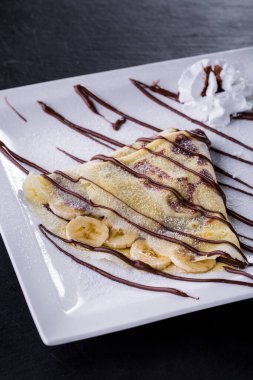 Dessert with bananas, pita bread, cream and chocolate topping on a white plate and stone background. Vertical orientation