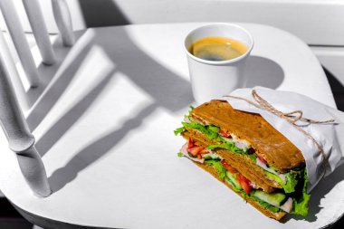 Sandwich in white paper with ham, lettuce, tomatoes, cucumbers and sauce with cappuccino with a paper cup standing on a white chair against the background of a white wooden wall