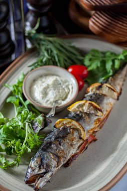 Baked stuffed grilled mackerel with lemon, frisee salad, parsley, cherry tomatoes, onion sauce in a saucepan on an oval plate. Vertical orientation 