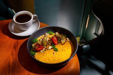 Pumpkin porridge with strawberries, apple, mint, avocado, sesame seeds, cheese and walnuts in a plate on the table with a cup of coffee