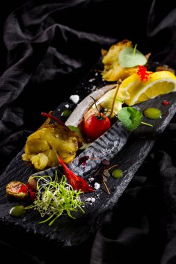 Patatesli fırınlanmış uskumru, limon dilimleri, çiçek yaprakları, tobiko havyarı, kumkuat, ve taş bir levha üzerinde taze filizler ve koyu bir arka plan. Dikey yönelim