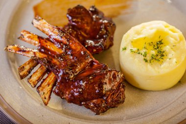 Grilled rack of lamb with barbecue sauce. Next to it is mashed potatoes with butter and dill. The food is on a round ceramic plate with sauce smeared on the plate. The plate is on a beige tablecloth, next to it is a wooden board with grilled toast, t