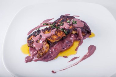 Kızarmış kaz ciğeri ezmesi, vişne soslu ananas ve kekik soslu. Tabağa biraz kiraz sosu bulaşmış. Kaz ciğeri beyaz bir terliğin üzerinde, tabak beyaz bir arka planda duruyor..