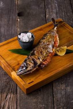 Grilled stuffed mackerel on a banana leaf with fried lemon and white sauce on a wooden board and wooden table. Vertical orientation