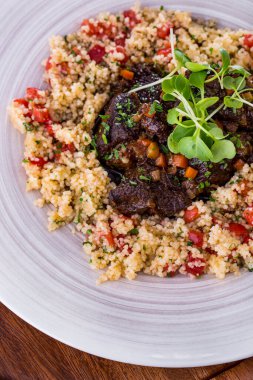 Couscous with tomatoes and chopped cilantro, stewed veal in a prune and carrot sauce. Sprinkled with chopped cilantro, pea microgreen sprouts on top. The food is in a white ceramic plate. The plate stands on a wooden background.