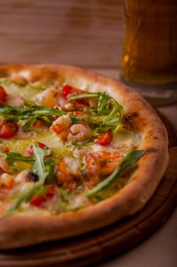Pizza with cherry tomatoes, shrimps, arugula and cheese on a wooden board with a glass of beer on the table