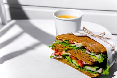 Sandwich in white paper with ham, lettuce, tomatoes, cucumbers and sauce with cappuccino with a paper cup standing on a white chair against the background of a white wooden wall